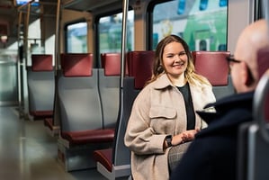 A woman smiles and sits on the train on her way to work. There is a man wearing glasses sitting across from her.