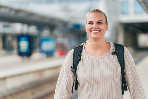 Hymyilevä nuori nainen juna-asemalla reppu selässään